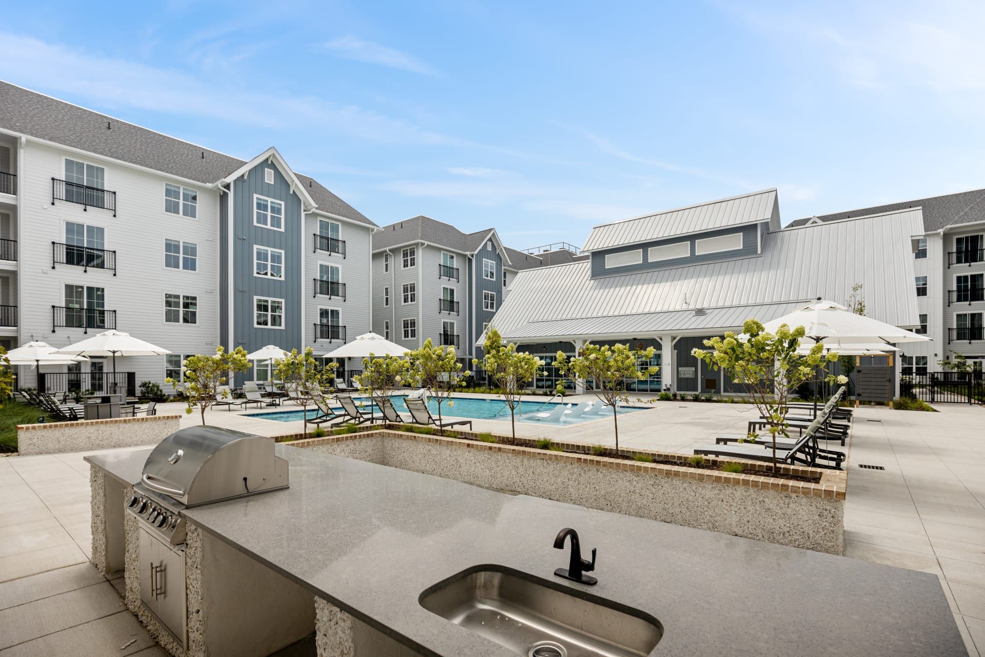 Outdoor kitchen area with a grill and sink overlooking a pool and lounge chairs at an apartment complex.