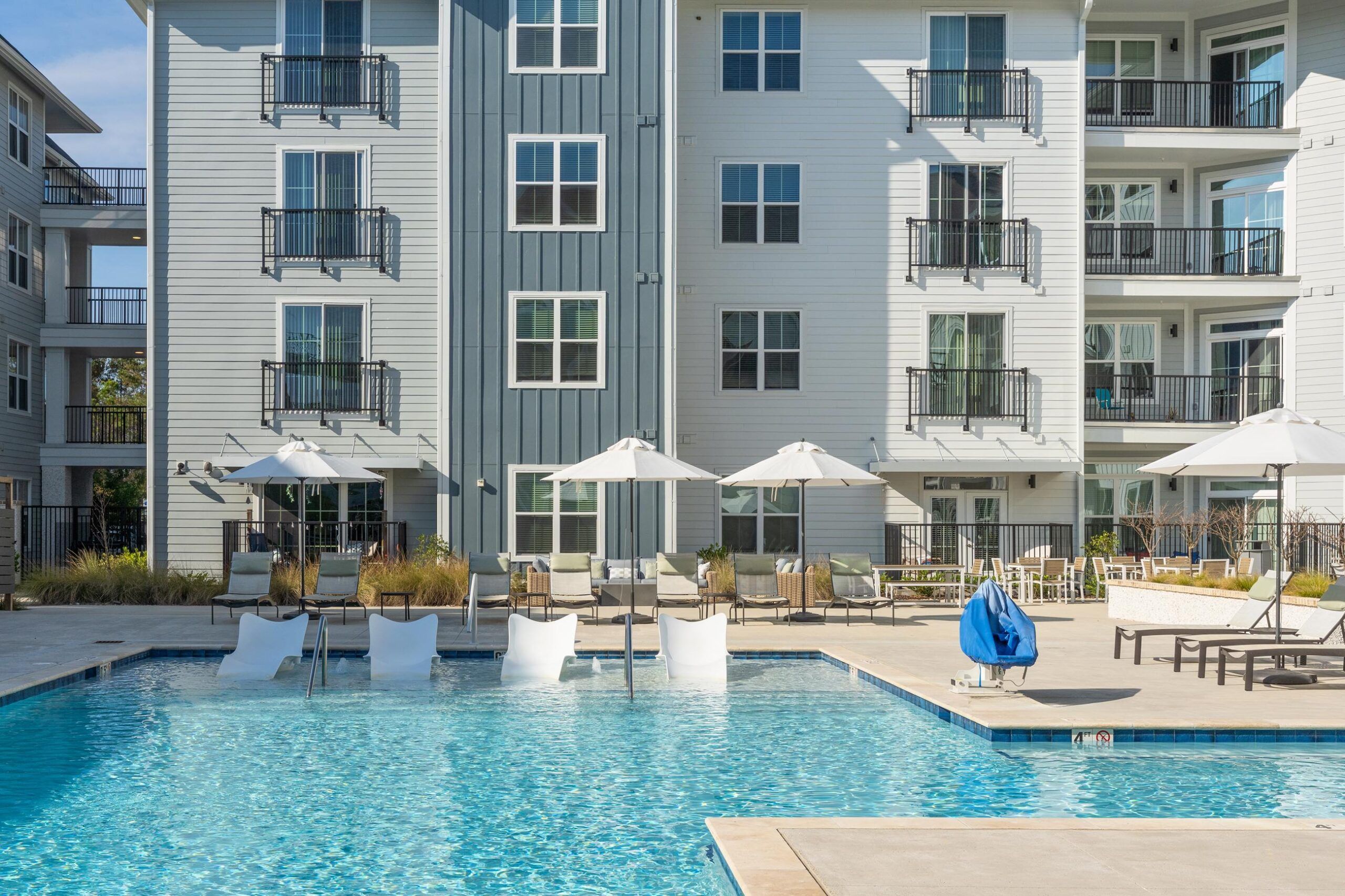 Modern apartment building with balconies overlooking a pool with lounge chairs and umbrellas.
