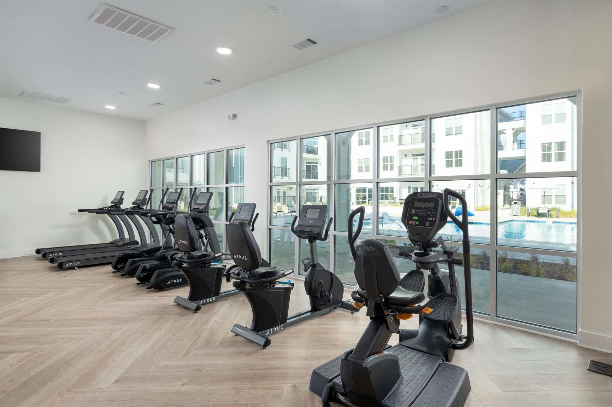Modern gym with treadmills, exercise bikes, and large windows overlooking a pool and apartment buildings.