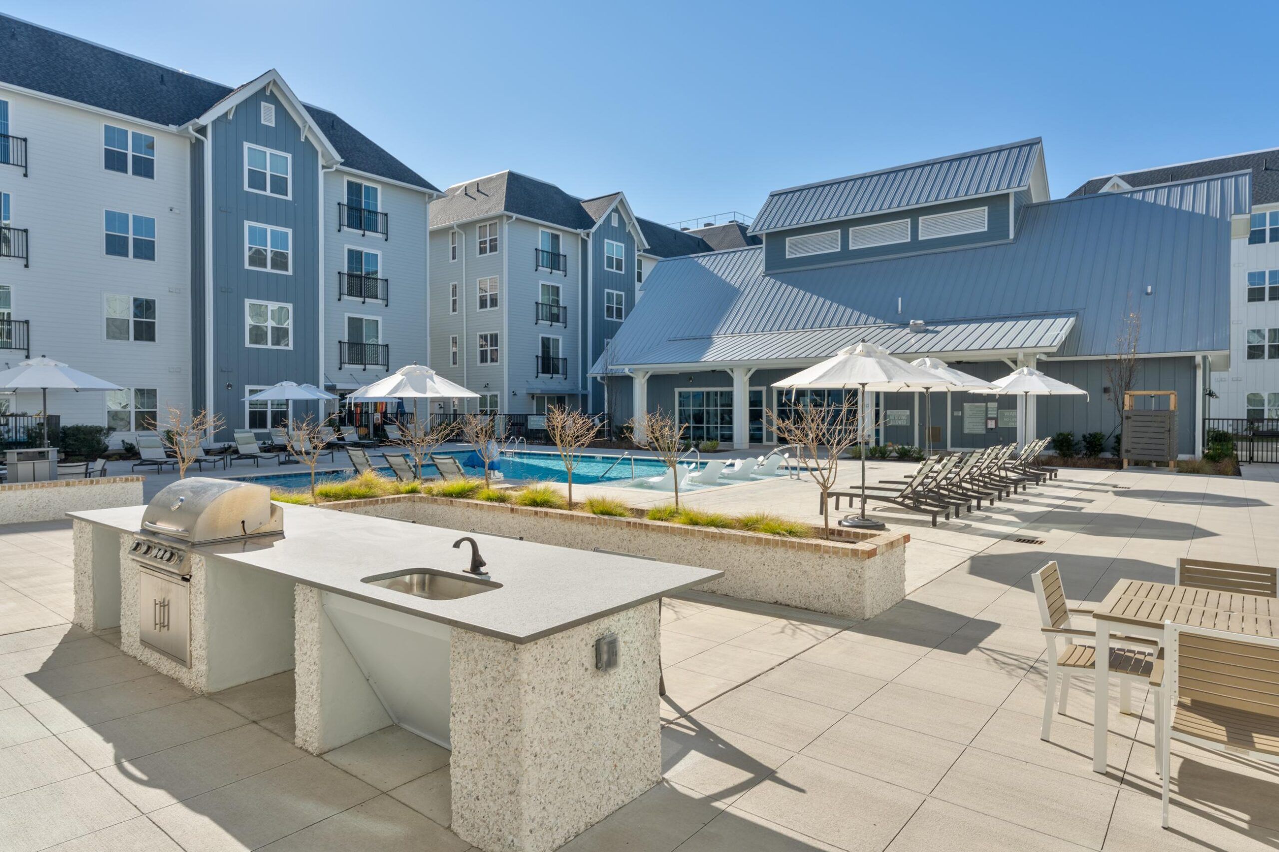 Modern apartment complex courtyard with pool, lounge chairs, umbrellas, and an outdoor kitchen with a grill.