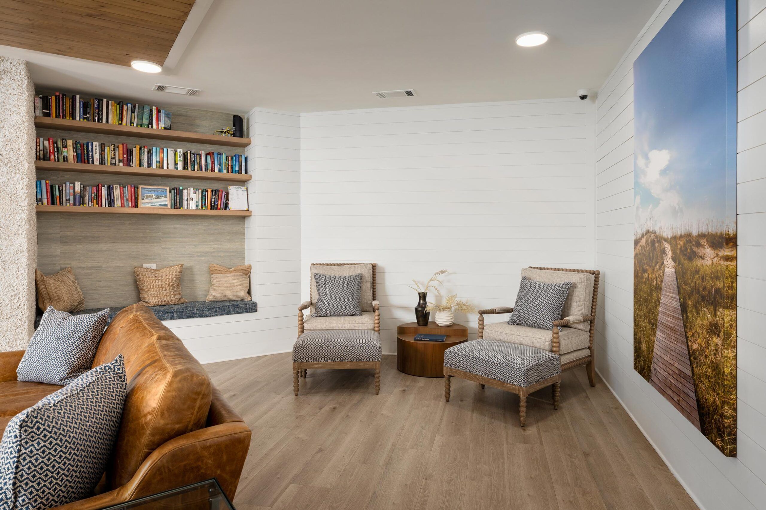 Cozy living room with armchairs, a leather sofa, bookshelves, and wall art of a boardwalk scene.